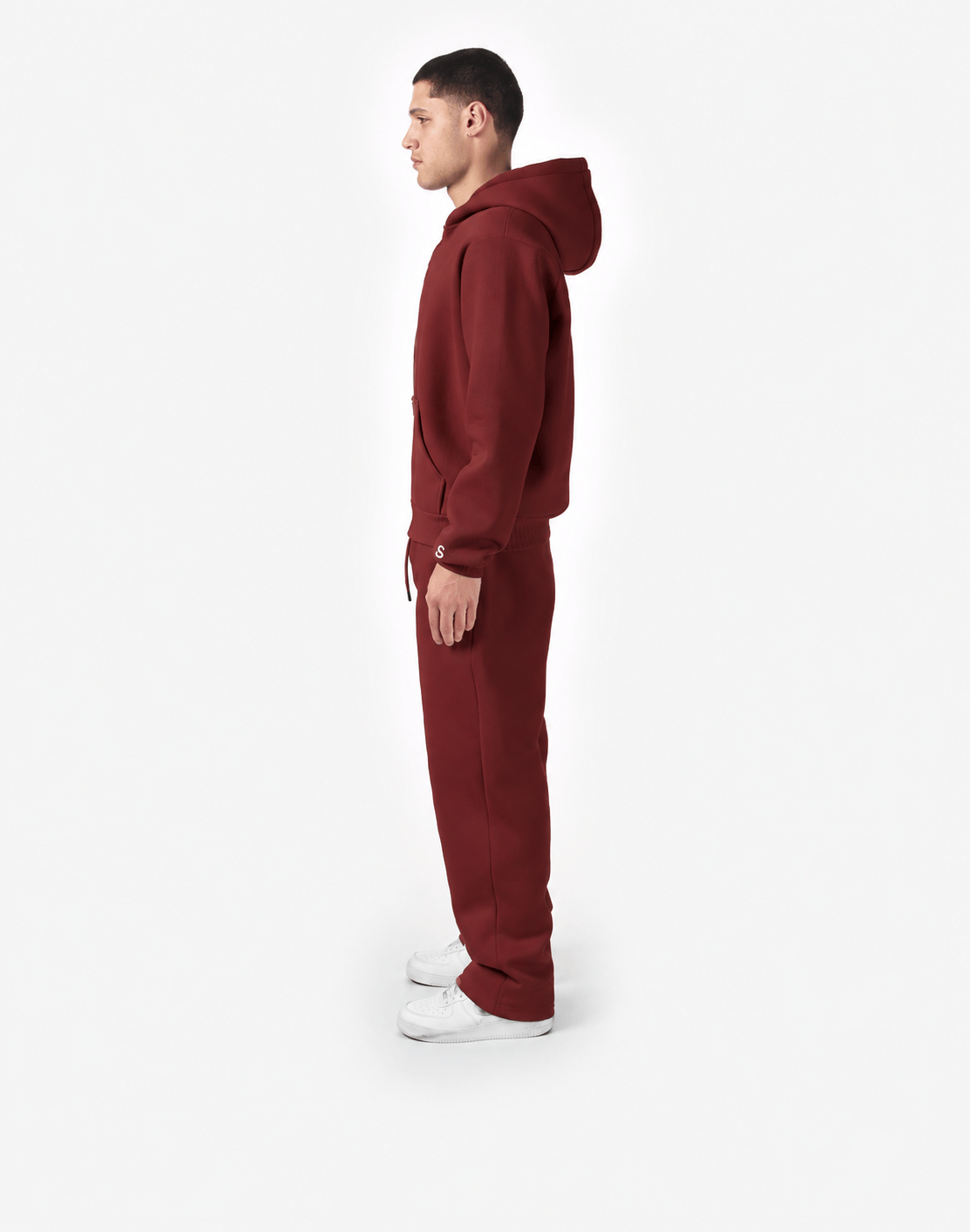 Model von hinten in einem bordeauxroten Trainingsanzug mit Kapuze und langer Hose, dazu weiße Sneaker vor hellem Hintergrund.